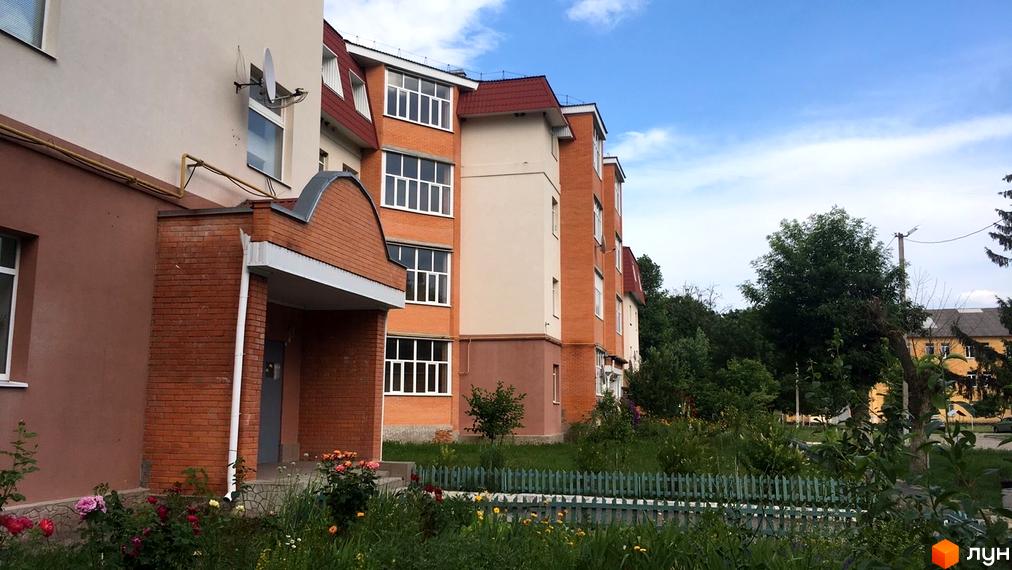 Зовнішній вигляд житлового комплексу на вул. Гайок, 224б, з цегляним фасадом та зеленими насадженнями навколо. Опис новобудови з квітами та парканом.