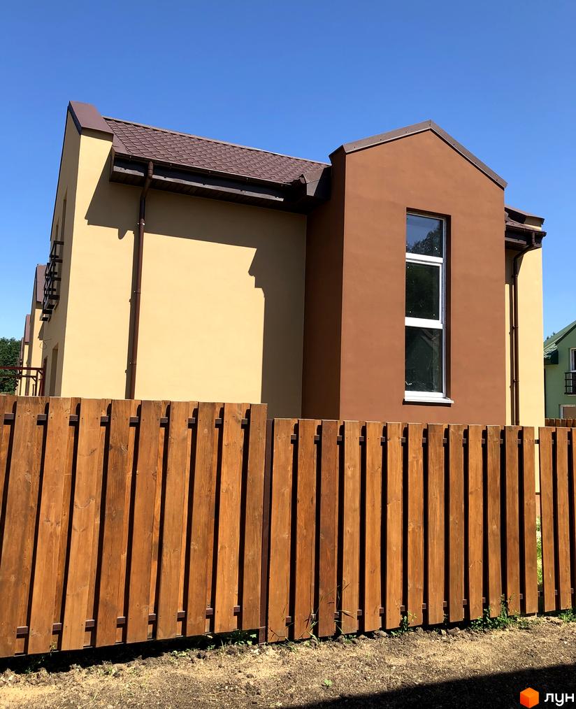 Будинок в Котеджному містечку Sunny Village має двокольоровий фасад: світло-бежевий та коричневий, з коричневим дахом. Перед будинком розташований дерев'яний паркан.