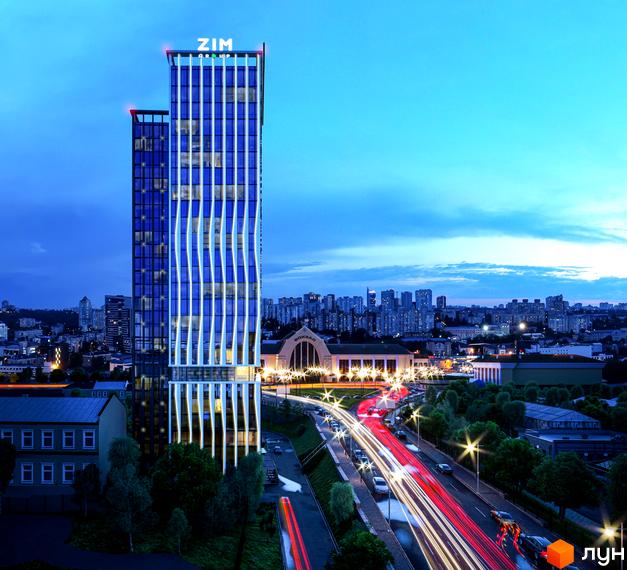 Апарт-комплекс Times ввечері: висотна будівля з синім склом та білими вертикальними елементами, поруч жвава дорога та міський вокзал.