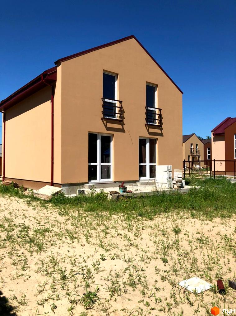 Котеджне містечко Sunny Village: двоповерховий будинок світло-коричневого кольору з червоним дахом та білими вікнами. Навколо піщана ділянка з рідкою травою.