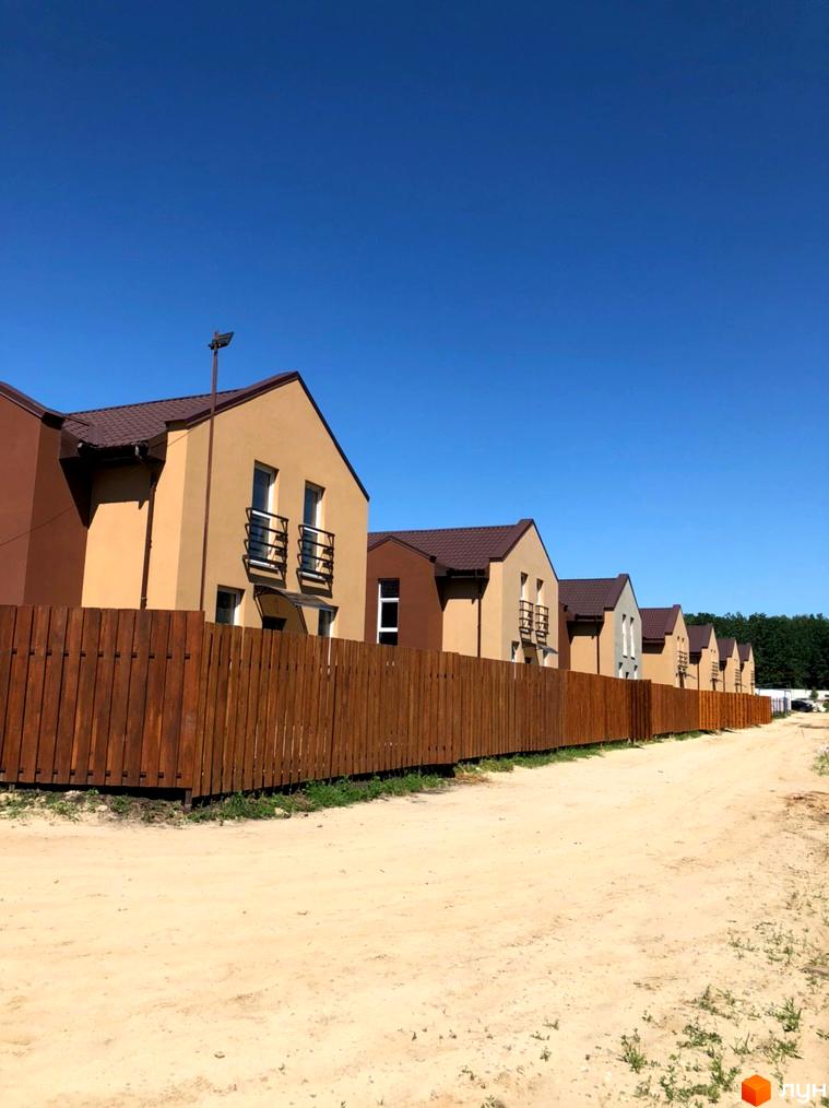 Котеджне містечко Sunny Village: ряд двоповерхових будинків з бежевими фасадами та коричневими дахами. Перед будинками розташований дерев'яний паркан та ґрунтова дорога.