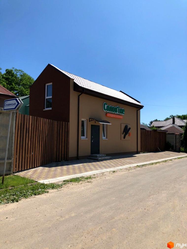 Будинок Котеджного містечка Sunny Village з бежевим фасадом, коричневим фронтоном та світлою черепицею. Поруч дерев'яний паркан, брукована доріжка та ґрунтова дорога.