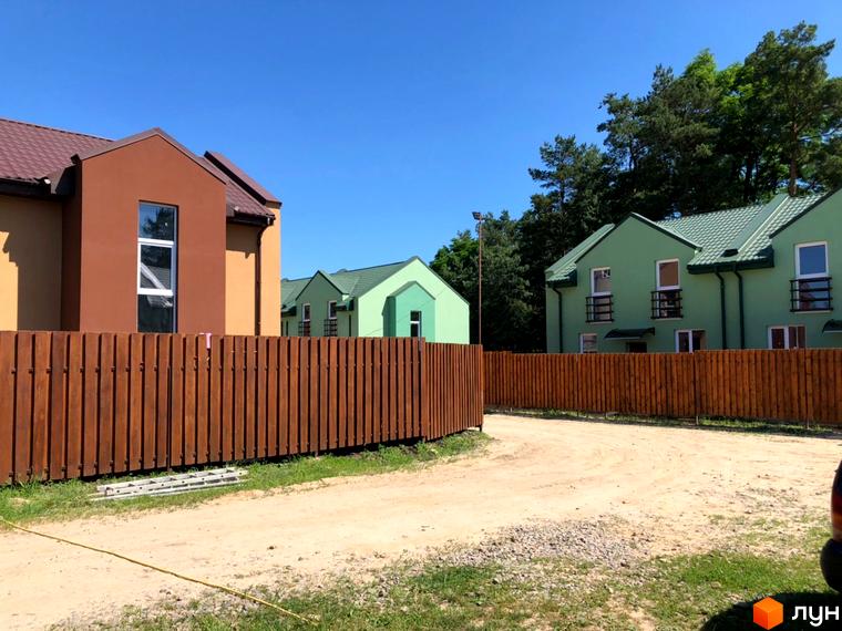 Будинки Котеджного містечка Sunny Village з коричневими, бежевими та зеленими фасадами, коричневими дахами, а також дерев'яний паркан і ґрунтова дорога.