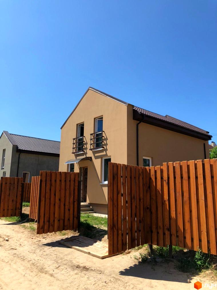 Будинки Котеджного містечка Sunny Village з фасадами бежевого та сірого кольорів, коричневими дахами. На передньому плані дерев'яний паркан та ґрунтова дорога.