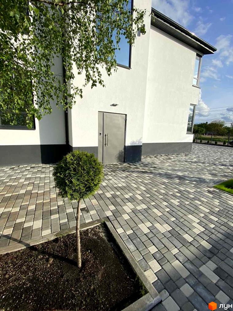 Новобудова ЖК Aura з білою фасадною штукатуркою та сірим цоколем, прибудинкова територія з бруківки та зеленої рослинності.