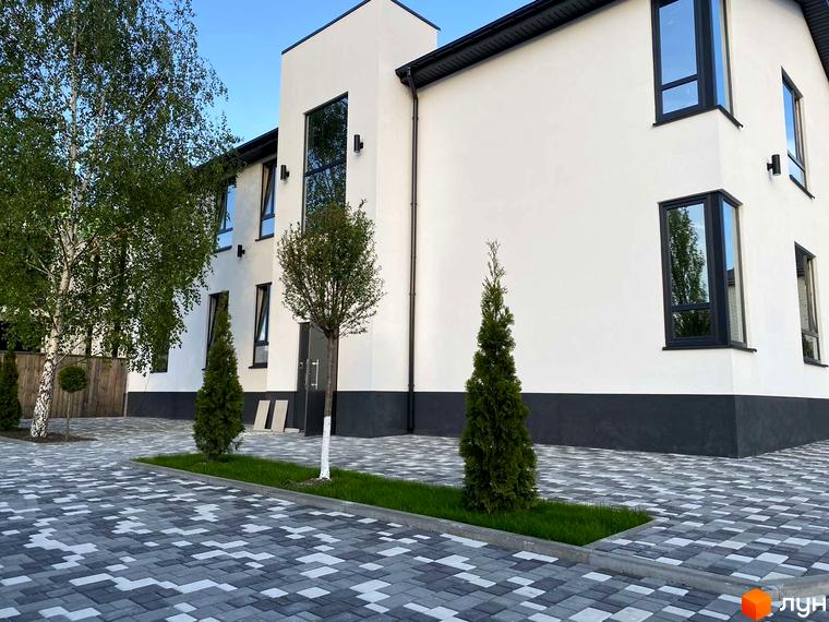 Фасад будинку житлового комплексу Aura з великими вікнами та сірою цокольною частиною. Прибудинкова територія вимощена плиткою з геометричним візерунком.