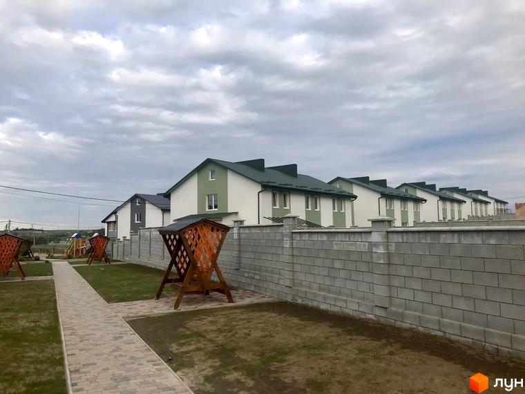 Зображення житлового комплексу таунхаусів. На фото видно ряд будинків з білими стінами та зеленими дахами, а також доглянуту прибудинкову територію з доріжками та альтанками.