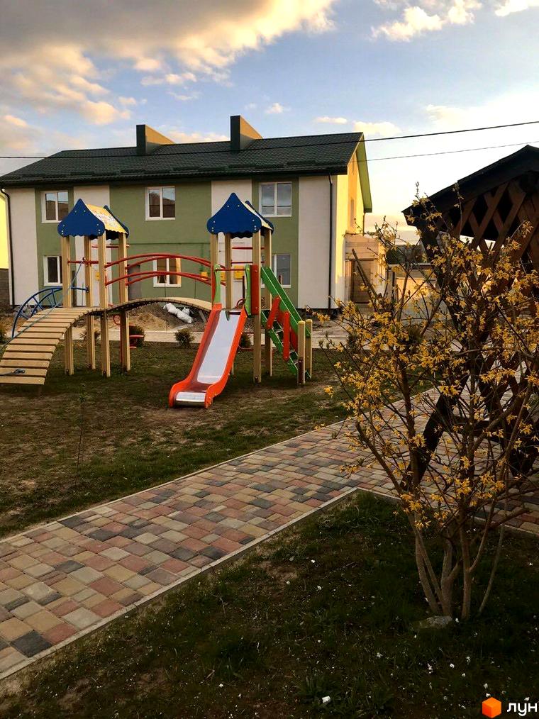 Житловий комплекс з дитячим майданчиком та доріжкою з бруківки. Будинок з зеленим дахом та білими стінами, поруч розташовані дерева.