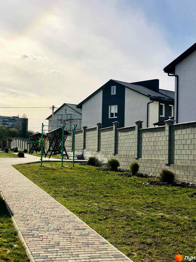 Зображення житлового будинку в Сонячній Долині. На передньому плані бруківка та газон, на задньому – паркан та будинки.