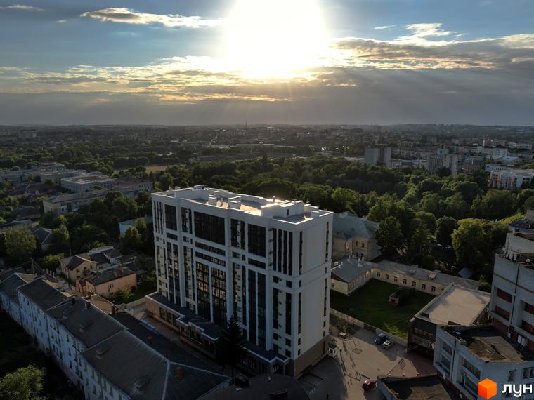 Висотна будівля ЖК Prestige Apartments на фоні неба та міста. Білосніжний фасад з темними вікнами, навколо зелені насадження та інші будівлі.