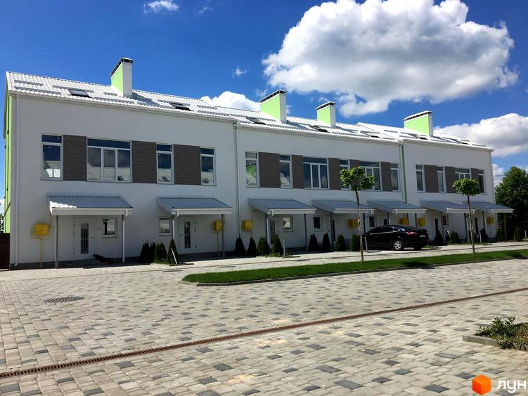 Ряд білих таунхаусів Таунхауси „Apple House“ з зеленими димарями та сірими дахами. Перед будинками вимощена бруківкою дорога та зелені насадження.