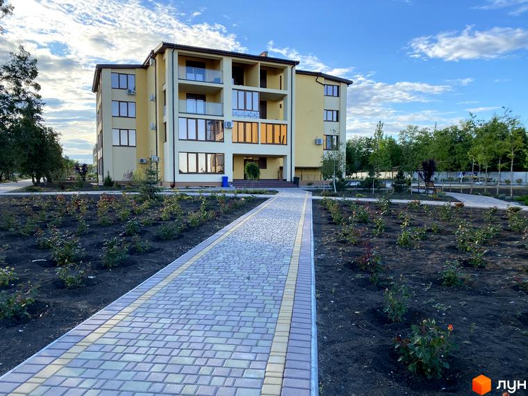 Багатоповерховий будинок GRAND FAMILY APARTMENTS світлого кольору з великими вікнами та балконами. Перед ним брукована доріжка та молоді насадження.