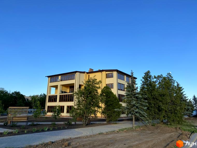 Односекційний будинок GRAND FAMILY APARTMENTS з бежевим фасадом та великими вікнами. Навколо будинку облаштована територія з доріжками та деревами.