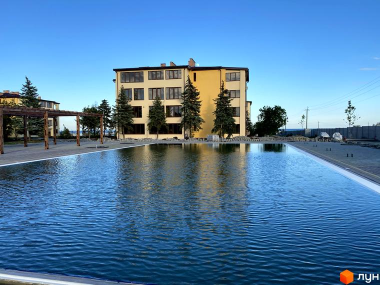 Будівля GRAND FAMILY APARTMENTS з кількома поверхами та світлим фасадом. Перед нею розташований великий басейн, а територія озеленена деревами.