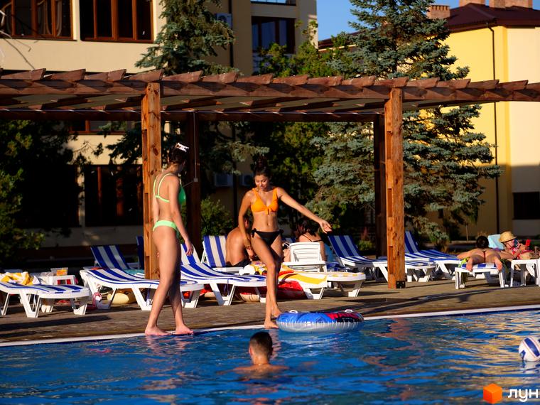 Багатоповерховий будинок GRAND FAMILY APARTMENTS світлого кольору з коричневими вікнами. На території є басейн та зона відпочинку з шезлонгами.