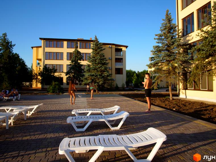 Будинки GRAND FAMILY APARTMENTS світло-жовтого кольору. На прибудинковій території бруківка, хвойні дерева та білі шезлонги.