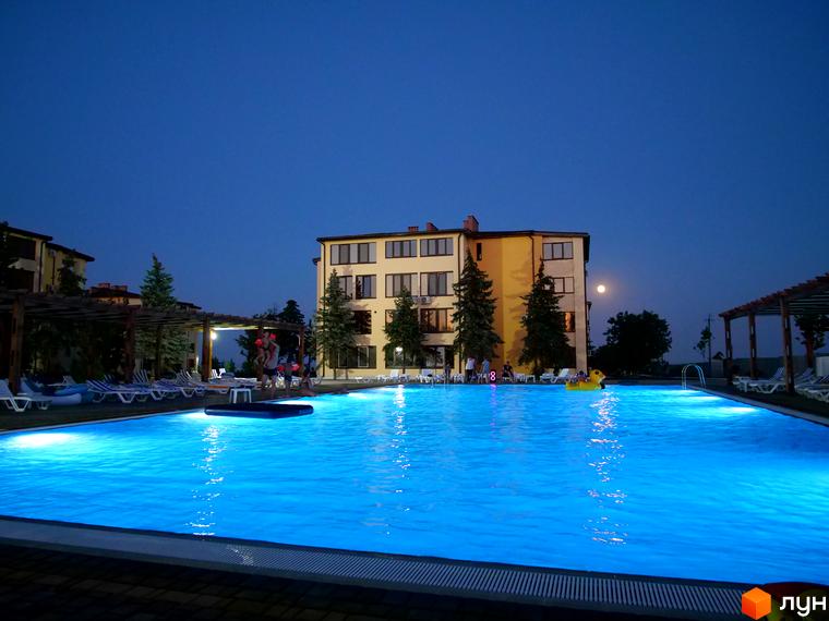 Будівля житлового комплексу GRAND FAMILY APARTMENTS з басейном та лежаками ввечері. Навколо будівлі ростуть дерева, а над басейном видно місяць.