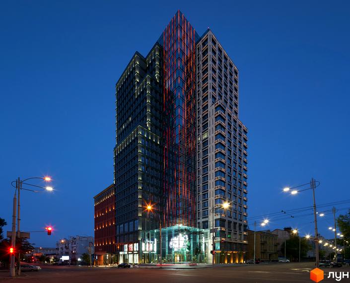 Висотна будівля ЖК Chicago Central House ввечері з підсвіткою. Навколо будівлі розташована міська інфраструктура: дороги, ліхтарі, будівлі.