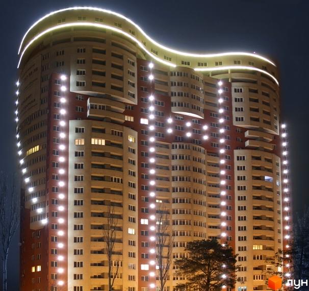 Житловий комплекс Воскресенський вночі з яскравою підсвіткою фасаду та хвилястим дахом. На передньому плані видно дерева без листя.