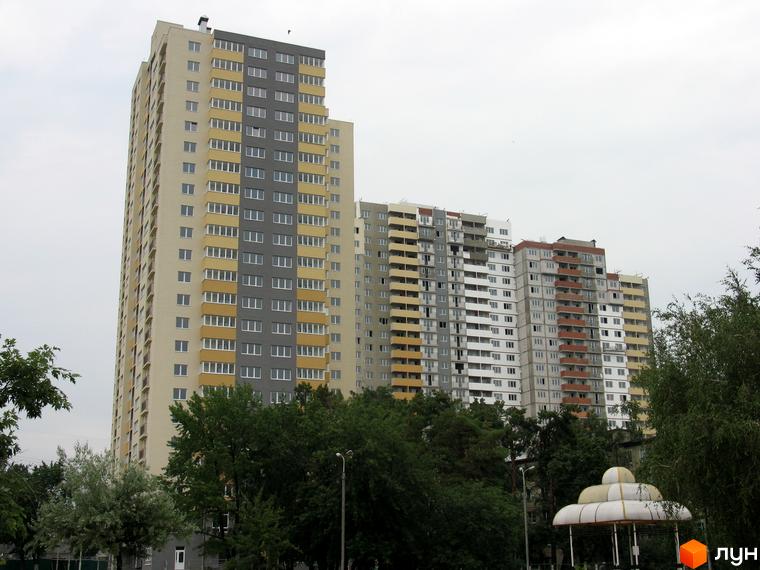 Зображення Житлового комплексу Перовський: високий житловий будинок з чергуванням бежевих та сірих секцій фасаду, балконами. Навколо видно зелені дерева та інші будівлі.