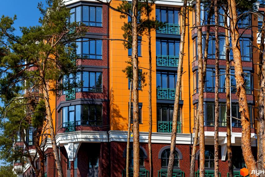 Клубний будинок „На Радистів“ з цегляним та помаранчевим фасадом, великими вікнами та зеленими балконами, оточений соснами.