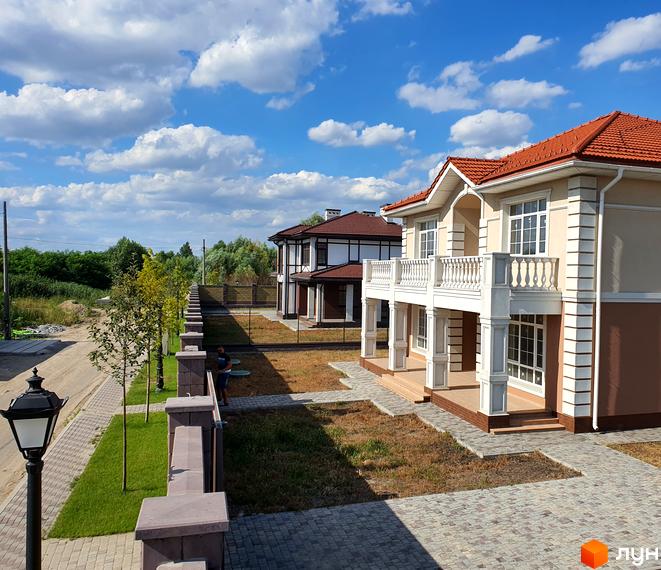 Двоповерховий будинок з червоним дахом та балконом у Котеджному містечку Осокор. На території є доглянутий газон та бруківка.