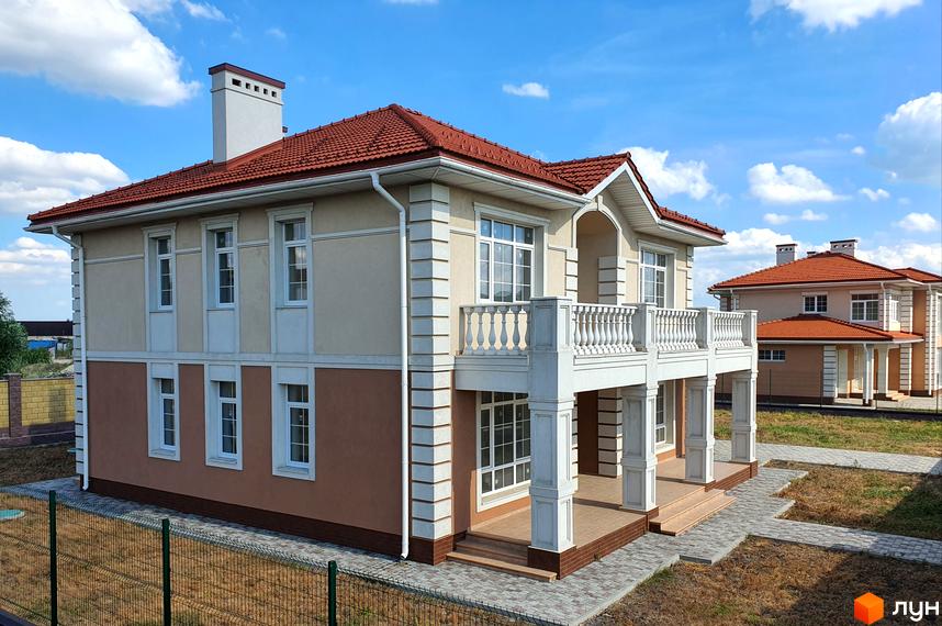 Двоповерховий будинок у Котеджному містечку Осокор з балконом та червоним дахом. Навколо будинку газон та тротуарна плитка.