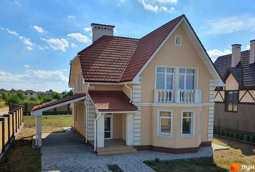 Двоповерховий будинок з коричневою черепичною покрівлею та балконом. Перед будинком викладена бруківка, є газон та зелені насадження.