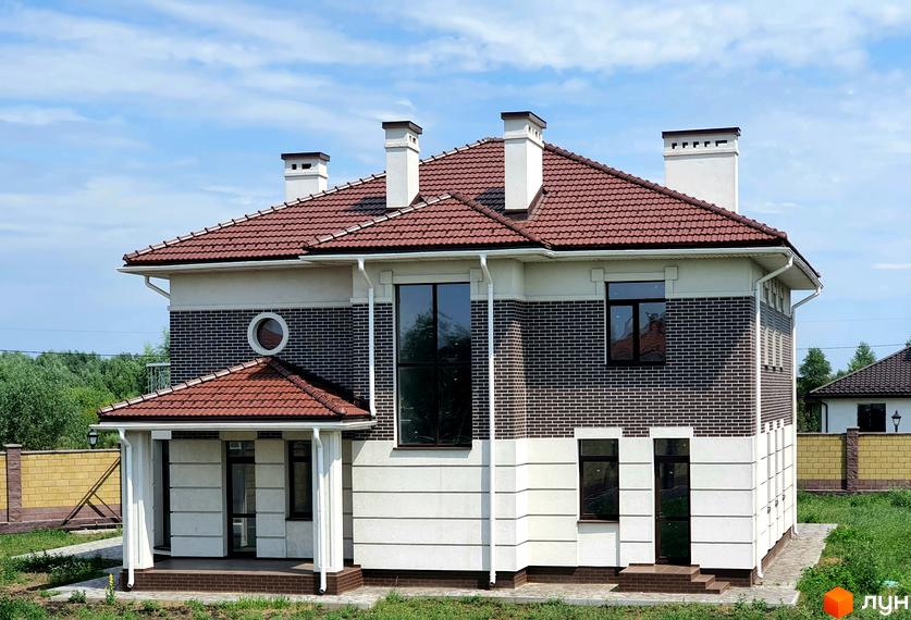 Двоповерховий будинок з темно-коричневою черепицею на даху та цегляною кладкою. На території зелена трава та огорожа з жовтої цегли.