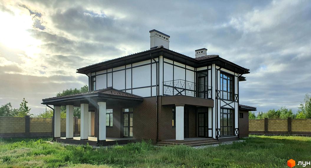 Двоповерховий будинок з білими стінами та коричневими балками, що створюють контрастний вигляд. Перед будинком зелена трава та огорожа з цегли.