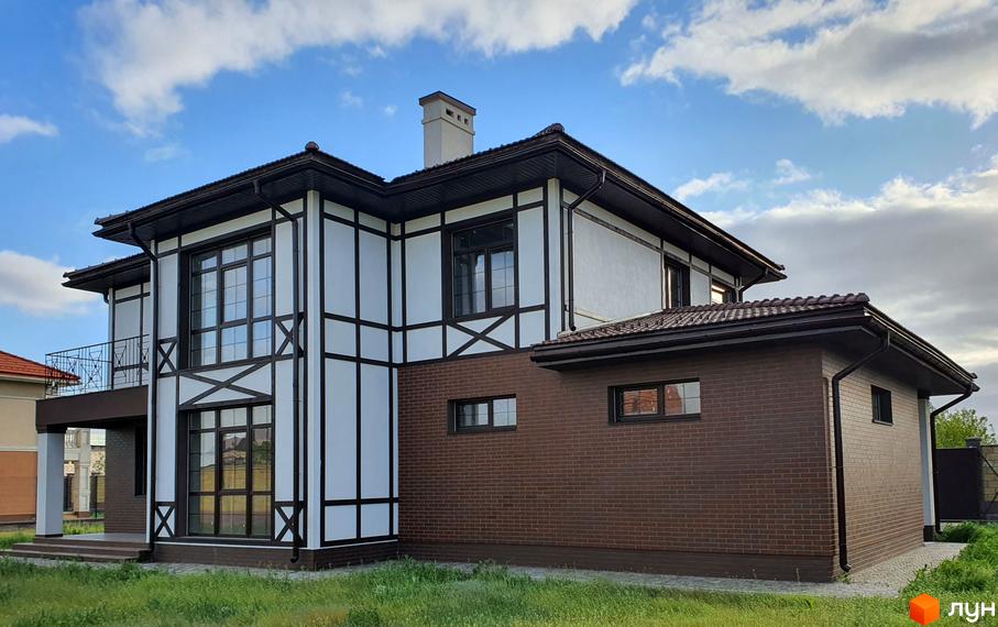 Двоповерховий будинок з білими стінами та темними балками, коричневою черепицею на даху. На території зелена трава та блакитне небо з хмарами.