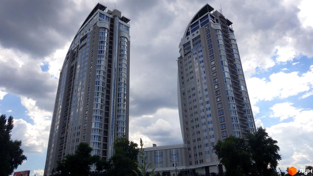 Дві високі житлові вежі світлого кольору з численними вікнами та балконами. Житловий комплекс на просп. Володимира Івасюка, 2, оточений зеленню під хмарним небом.