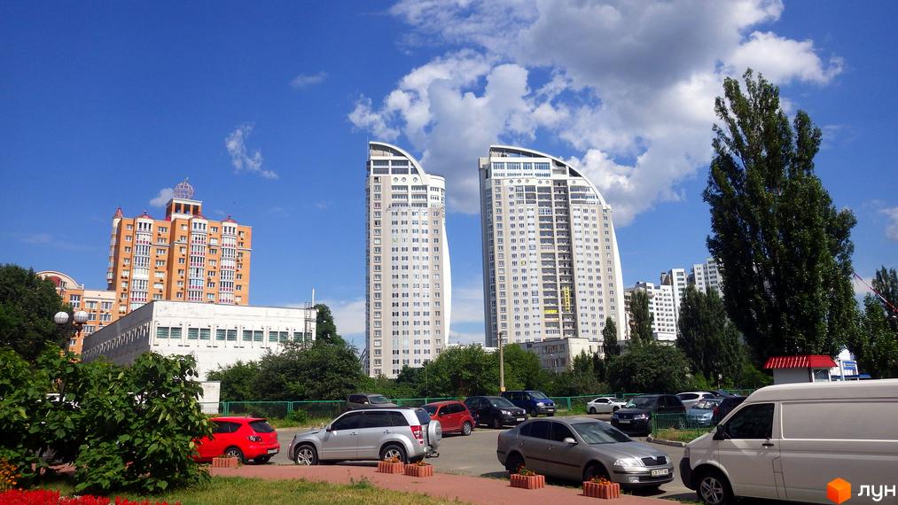 Вид на Житловий комплекс за адресою просп. Володимира Івасюка, 2, з двома високими світлими будинками та прилеглою територією з парковкою та зеленими насадженнями.