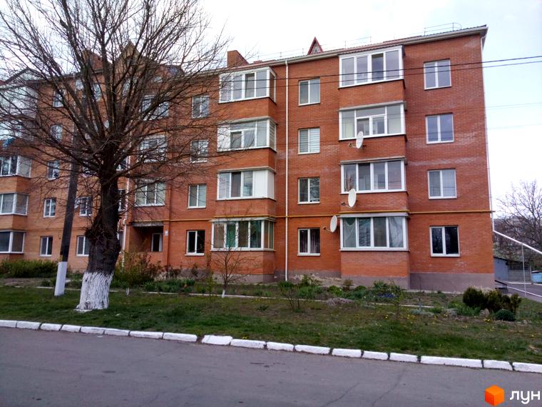 Зовнішній вигляд цегляної новобудови з численними вікнами та балконами, що розташована на фоні зеленої прибудинкової території.