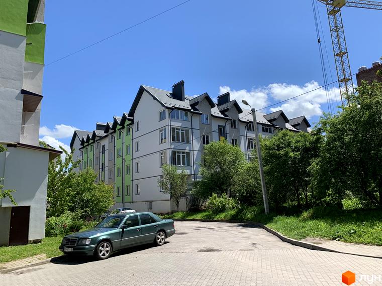 Зовнішній вигляд новобудови ЖК Мілан з зеленими та білими фасадами, що розташована біля дороги з прибудинковою територією та деревами. На передньому плані зелений автомобіль.