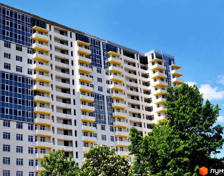 Житловий комплекс Перлина Нивок з білим фасадом, жовтими балконами та великими вікнами. На передньому плані зелені дерева.