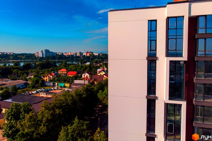 Житловий район Manhattan: сучасна архітектура з великими вікнами та цегляними вставками на тлі зеленого міського пейзажу з річкою та малоповерховою забудовою.