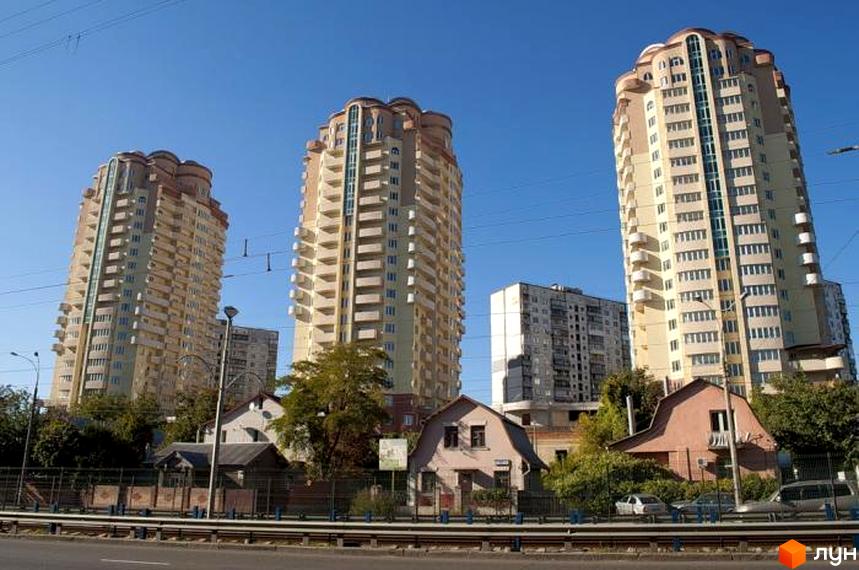 Три високі світлі будинки Житлового комплексу Три Богатирі з багатьма балконами та округлими дахами. На передньому плані видно приватні будинки, дерева та дорогу.