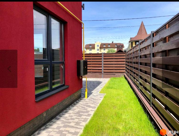 Таунхауси Old Stories з червоною стіною та вікном, огороджені дерев'яним парканом. Прибудинкова територія з газоном та бруківкою.