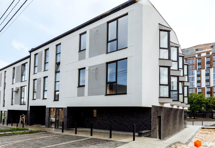 Клубний будинок Craft - це сучасна новобудова з панорамними вікнами та сірими акцентами на фасаді. Перед будинком розташований вимощений паркінг та зелені насадження.