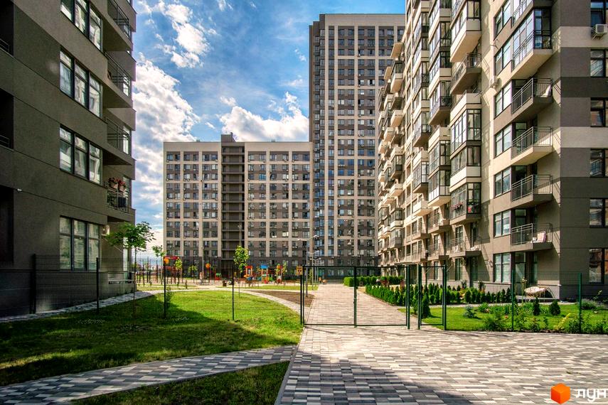Зовнішній вигляд житлового комплексу Варшавський Мікрорайон з високими будинками та балконами. На прибудинковій території розташований дитячий майданчик та зелені насадження.