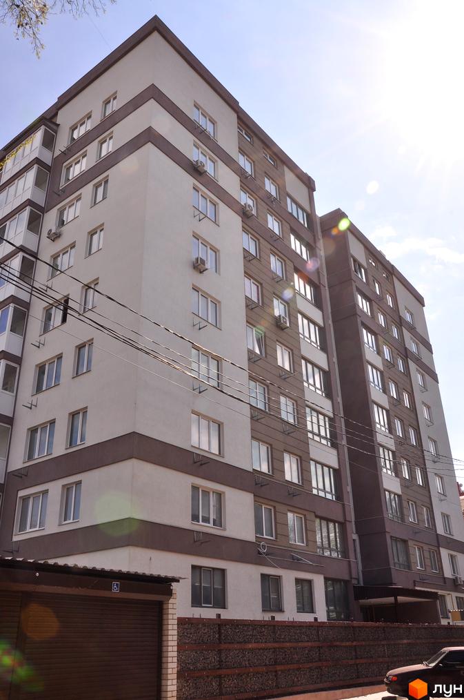 Зовнішній вигляд новобудови ЖК Мирний: багатоповерховий будинок з біло-коричневим фасадом та заскленими балконами. Прибудинкова територія з парканом та гаражними воротами.