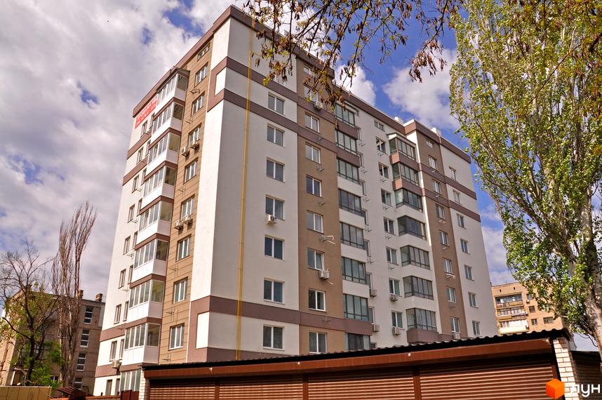 Зовнішній вигляд новобудови ЖК Мирний з білими та коричневими фасадами, скляними балконами та кондиціонерами. Навколо будинку видно дерева та хмарне небо.