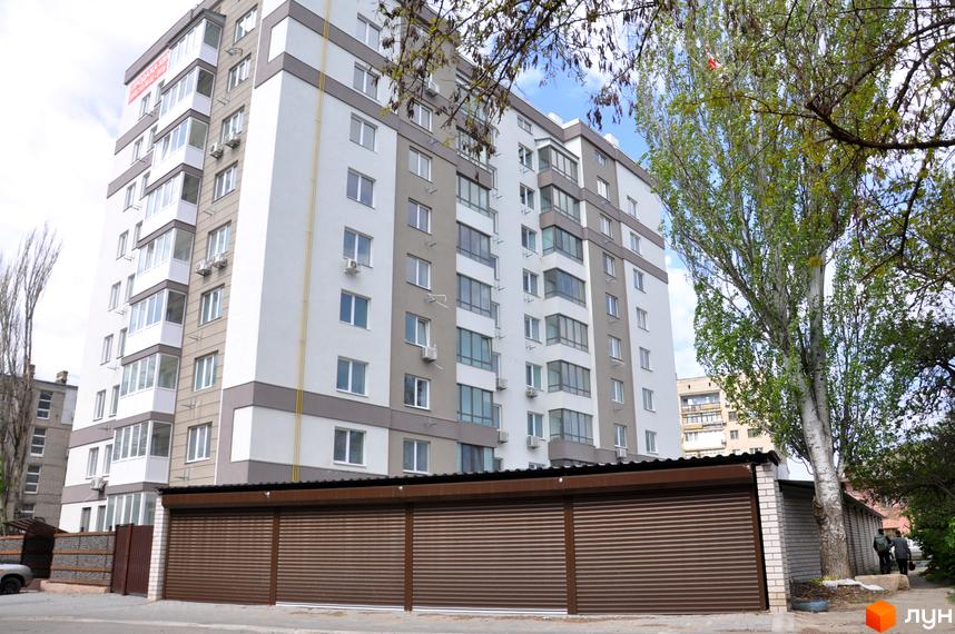 Зовнішній вигляд новобудови ЖК Мирний з коричневими ролетами та парканом. Навколо будівлі зелені дерева та частина сусіднього будинку.