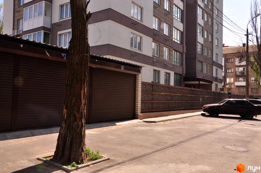 Зовнішній вигляд новобудови ЖК Мирний з коричневими ролетами та парканом з каменю. Прибудинкова територія з деревом та автомобілем.