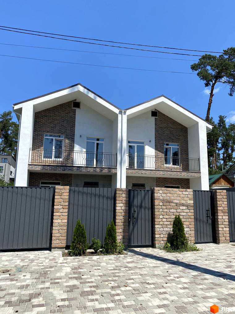 Двоповерховий дуплекс з цегляним фасадом та балконами, огороджений парканом з металопрофілю та цегли. Прибудинкова територія з бруківкою та туями.