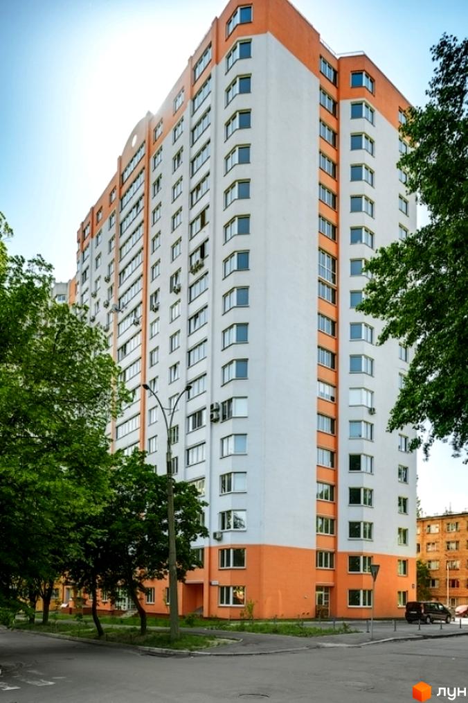 Багатоповерховий житловий будинок біло-помаранчевого кольору з численними вікнами, розташований біля дороги та оточений зеленими деревами.