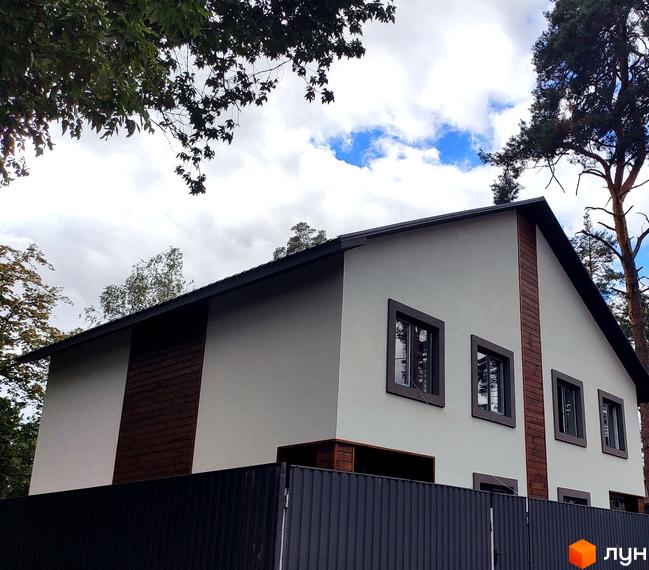 Зовнішній вигляд двоповерхового будинку з темним дахом та вікнами, оздоблений дерев'яними панелями. Навколо будинку видно зелені дерева та паркан.