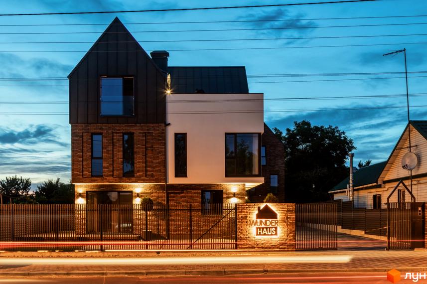 Котеджне містечко Wunder Haus з цегляним фасадом та темним дахом, освітлене ввечері. Огороджена територія з підсвіченим написом та доглянутими деревами.