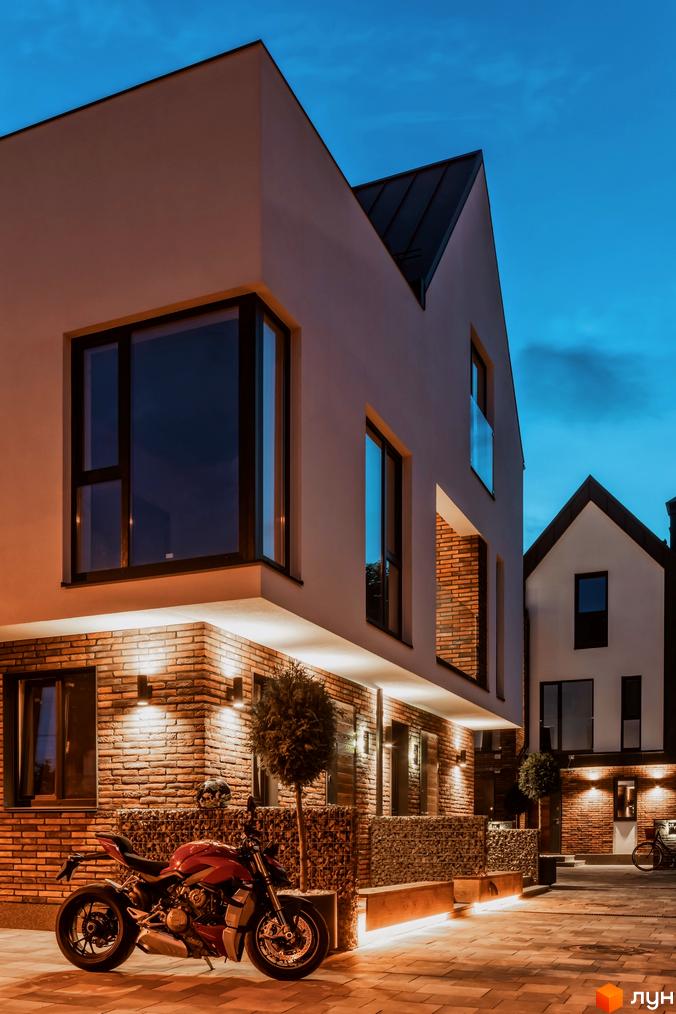 Котеджне містечко КМ Wunder Haus з цегляним фасадом та великими вікнами ввечері. На прибудинковій території розташований мотоцикл та лавки.